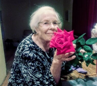 A woman in a black and pale green print, with gray hair and glasses, displays a blooming dark pink rose.