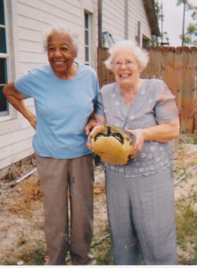 Two older women stand in what appears to be the back or side yard of a white wooden house, with a fence behind them. The woman on the left is African-American and wears a light blue shirt, brown pants, and glasses. The woman on the right is in a gray shirt with some pink in the sleeves and white dots on the torso. She wears gray pants and holds a large turtle.