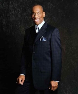 A professional photo shows a man dressed in a navy suit standing against a gray backdrop and smiling.